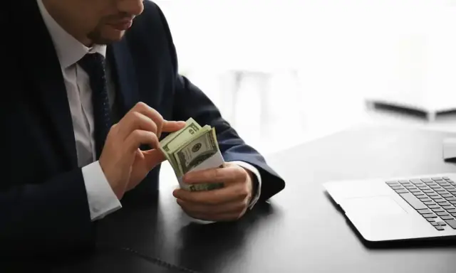 Mężczyzna w garniturze liczy pieniądze, zastanawiając się, ile zarabia osoba pracująca w banku. Obok leży laptop.