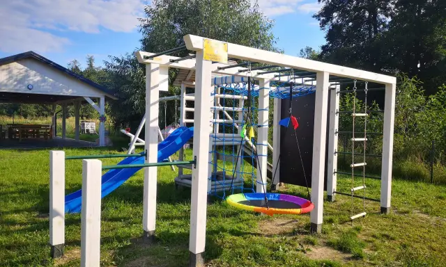 Plac zabaw w **dolinie roztoczy**: zjeżdżalnia, drabinki, huśtawka i ścianka wspinaczkowa. W tle altana.