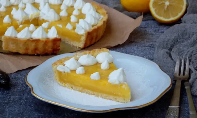 Une part de tarte au citron, garnie de meringue, sur une assiette. La tarte entière est visible en arrière-plan. Une recette de tarte au citron parfaite pour le dessert.