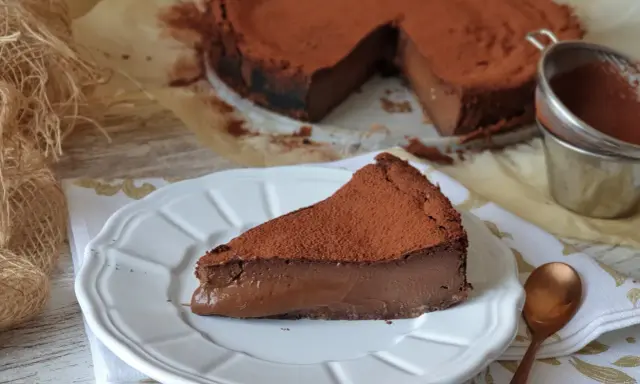 Una porción de tarta de chocolate, cubierta de cacao en polvo, reposa en un plato blanco. El resto de la tarta, también espolvoreada, se ve al fondo.
