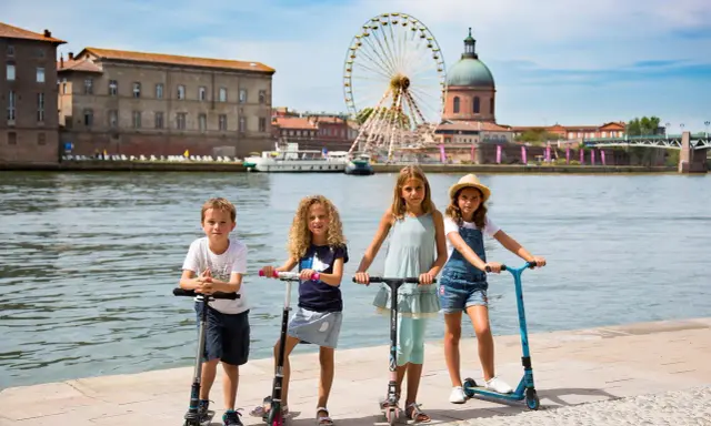 Niños en patinetes junto al río en Toulouse, con la noria y la cúpula de la Basílica de San Sernín de fondo. ¡Un plan genial que hacer en Toulouse con niños!