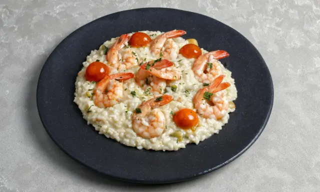 Kremowe risotto z krewetkami, pomidorkami koktajlowymi i posiekaną natką pietruszki na czarnym talerzu.