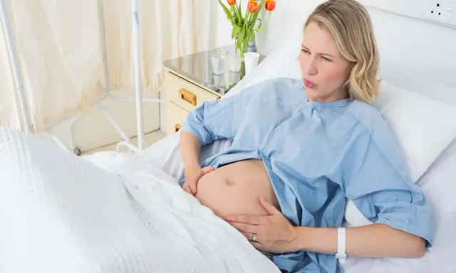 Gravid kvinna i sjukhussäng, med händerna på magen, ser ut att ha värkar. Vattnet har gått och hon väntar på barnet.