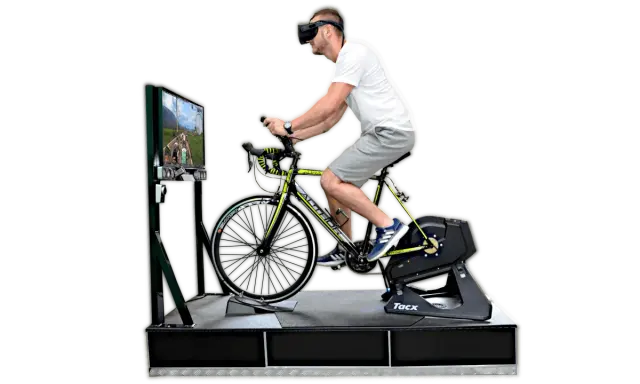 Mężczyzna w goglach VR pedałuje na rowerze stacjonarnym, doświadczając wirtualnej przejażdżki. To zaawansowany symulator jazdy rowerem.