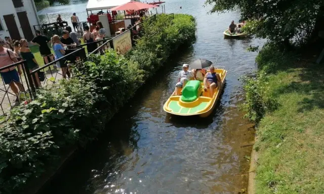 Rodzina pływa rowerkiem wodnym po kanale przy łagowskim jeziorze. Słońce odbija się w wodzie, a na brzegu widać ludzi odpoczywających w cieniu.