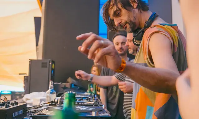 DJ Villa Lobos, con cuffie al collo, mixa musica con passione. Dietro di lui, il pubblico attende il ritmo.