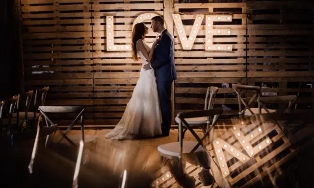 Para nietypowe sesje ślubne, para młoda pozuje przed podświetlanym napisem "LOVE" na tle drewnianych palet.