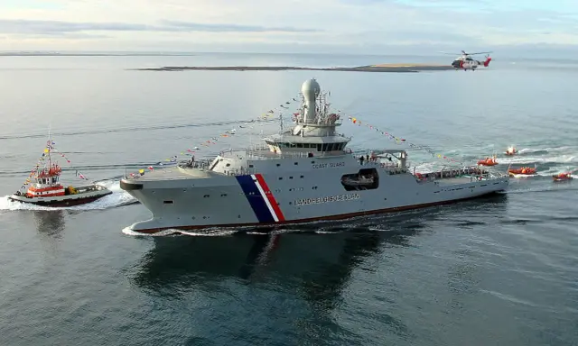 Un imposant bateau garde côte, le Landhelgisgæslan, navigue en mer, escorté par un remorqueur et suivi par des canots. Un hélicoptère survole la scène.