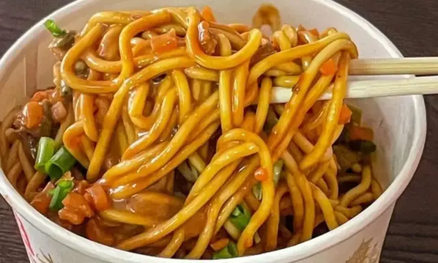 Un tazón lleno de fideos chinos salteados con verduras y salsa. Palillos levantan un bocado.