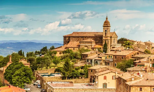 Panoramica del centro storico di Montalcino, con la chiesa e le case in pietra sotto un cielo azzurro con nuvole.