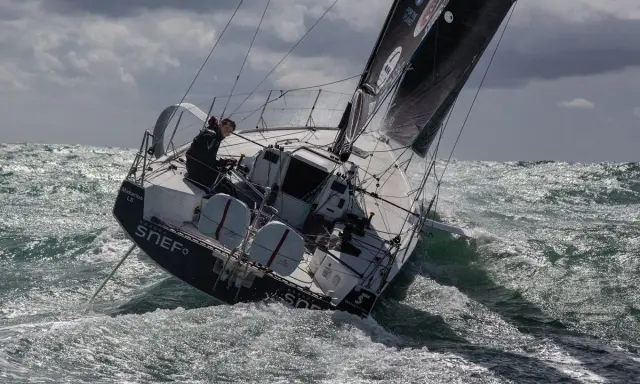 Xavier Macaire navigue sur un voilier dans une mer agitée, le bateau est incliné, les vagues déferlent.