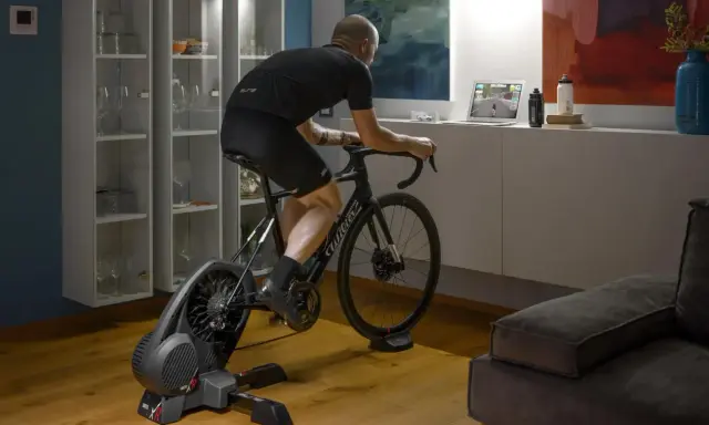 Mężczyzna trenuje na rowerze stacjonarnym z napędem **direct drive**, śledząc wirtualną trasę na laptopie.