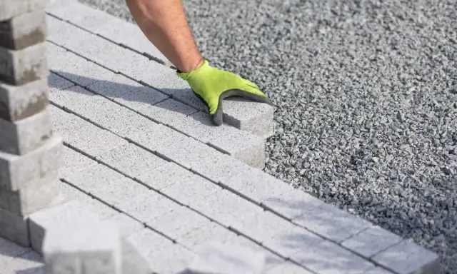 Ręka w zielonej rękawicy układa kostkę brukową na podsypce cementowo piaskowej.