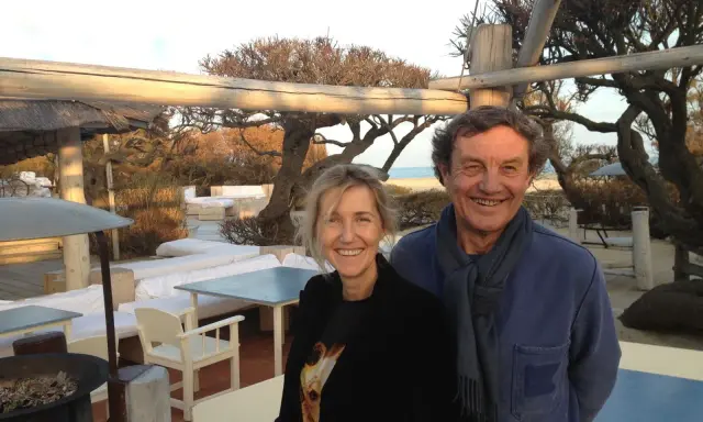 Patrice de Colmont et une femme souriants sur une terrasse de plage, avec des arbres dénudés et des sièges blancs en arrière-plan.