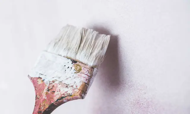 Pincel desgastado con restos de pintura blanca a punto de tocar una pared de cal con textura sutil.