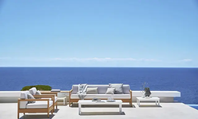 Un elegante divano Bali in legno e cuscini chiari, con vista sul mare blu intenso sotto un cielo limpido.