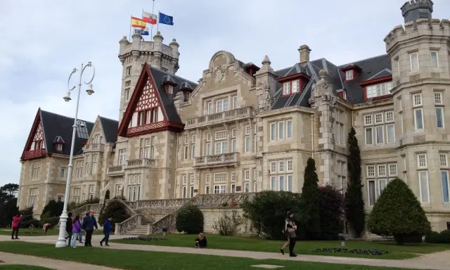Palacio de la Magdalena en Santander, con banderas ondeando en la torre. Turistas pasean por los jardines de esta joya arquitectónica.