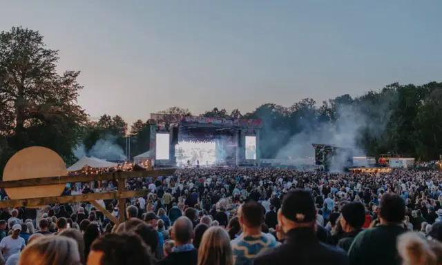 Stor publik vid Rosendal Garden Party. En stor scen med artister och ljus. Rök och träd i bakgrunden.