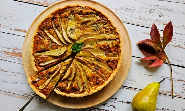 Apetyczna tarta z gruszkami, udekorowana natką pietruszki i skórką cytrynową, prezentuje się na drewnianej desce. Obok leży świeża gruszka i jesienne liście.