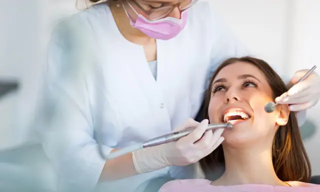 Dentystka w masce i rękawiczkach bada zęby pacjentki za pomocą lusterka i wiertła.