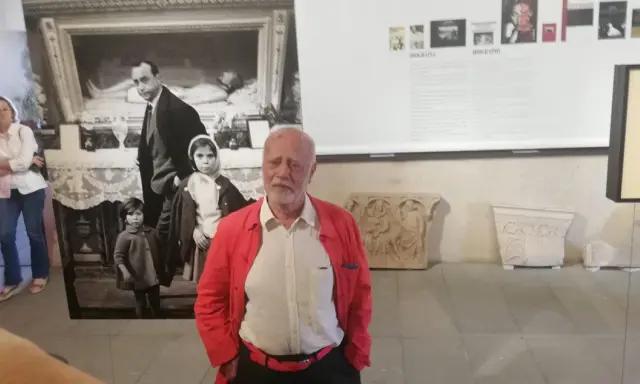 Un uomo anziano con giacca rossa, davanti a una foto di Ferdinando Scianna in Sicilia con bambini.