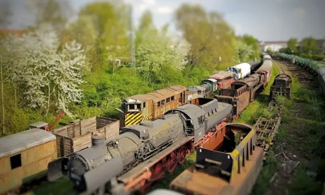 Un cementerio de trenes con efecto tilt shift, donde locomotoras y vagones oxidados parecen miniaturas bajo un cielo nublado.