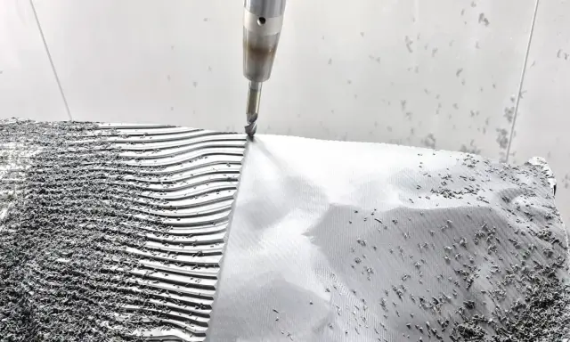 CNC-Fräser bearbeitet eine **Aluminiumstange**, die in Wellen und Rillen geformt wird. Späne bedecken die Oberfläche.