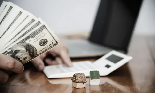Ręka trzyma banknoty 100 USD, obok kalkulator, laptop i miniaturowe domki. Zastanawiasz się, ile wynosi zasiłek socjalny na mieszkanie?