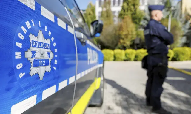 Policjant w mundurze stoi obok radiowozu z widocznym znakiem Policji i numerem 112.