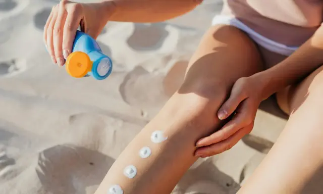 Main sur le sable, une main applique des points de crème solaire sur une jambe. Attention, cette crème solaire perimee pourrait ne plus protéger efficacement.