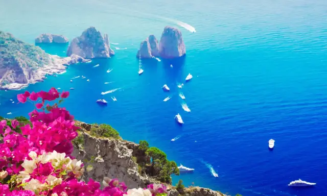 Un giorno a Capri: scogliere rocciose, mare blu intenso e barche che solcano le onde, con bouganville rosa in primo piano.