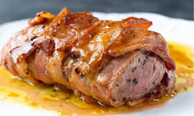 Saftiges Schweinefilet im Speckmantel, glasiert und mit Pfeffer bestreut, ruht in einer köstlichen Sauce auf einem weißen Teller.
