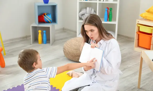 Terapeuta pracuje z dzieckiem nad poprawą niewyraźnej mowy, wskazując palcem na strony otwartej książki.