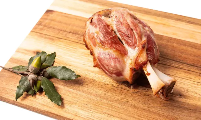 Codillo de cerdo asado, jugoso y dorado, reposa sobre una tabla de madera junto a unas bellotas.