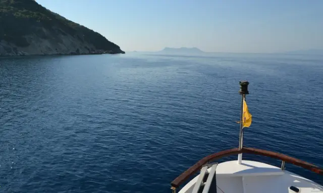 La prua di una barca naviga verso un'isola lontana, un viaggio che evoca la poesia di Itaca.