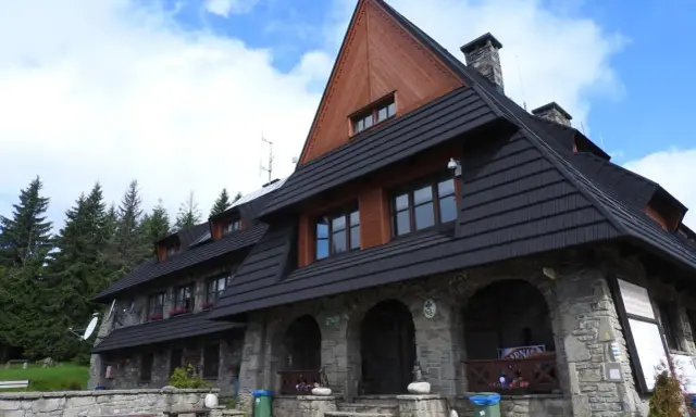 Kamienny schron turystyczny pod gorącymi szczytami, z drewnianą przybudówką i czarnym dachem, otoczony lasem świerkowym.