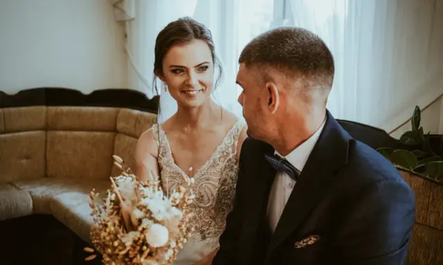Uśmiechnięta panna młoda w sukni z koronkami i pan młody w granatowym garniturze. W tle kanapa i zasłony. Krótka ceremonia, bo ile trwa ślub cywilny?