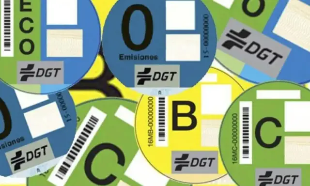 Varias etiquetas DGT circulares de diferentes colores (azul, verde, amarillo) con letras (ECO, 0, B, C) y códigos de barras, indicando la clasificación ambiental de vehículos.