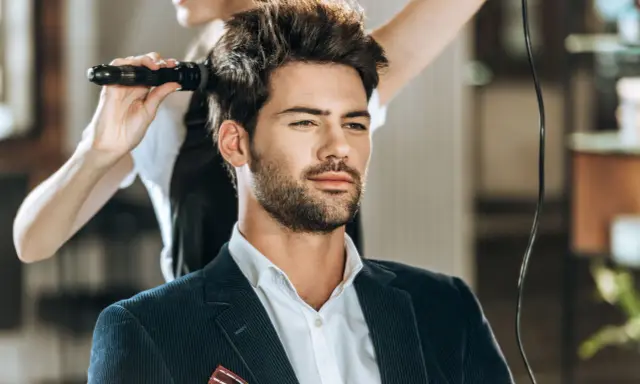 Mężczyzna w eleganckim garniturze czeka na stylizację fryzury męskiej. Fryzjerka suszy jego włosy suszarką.