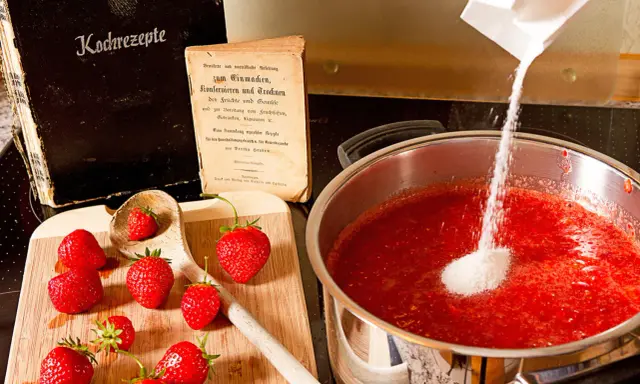 Zucker wird zu Erdbeeren in einem Topf gegeben, um zu erfahren, wann wird Marmelade fest. Alte Kochbücher liegen daneben.