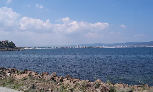 Słoneczny brzeg jak pić. Widok na morze i miasto w oddali, z kamienistym wybrzeżem na pierwszym planie.