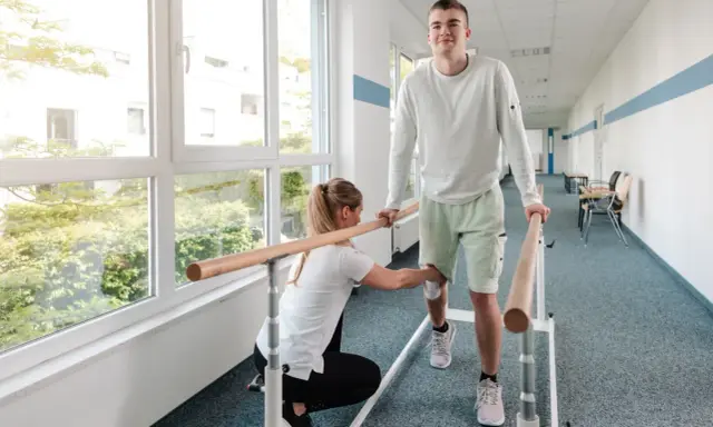 Młody mężczyzna ćwiczy chód przy poręczy, a fizjoterapeutka pomaga mu w rehabilitacji.