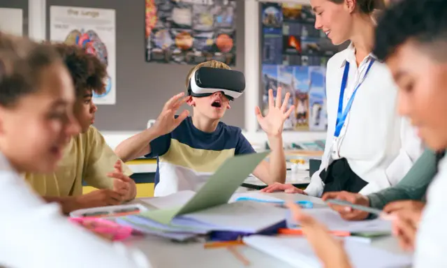 Chłopiec w goglach VR przeżywa lekcję, a nauczycielka z uśmiechem obserwuje tę innowację pedagogiczną.