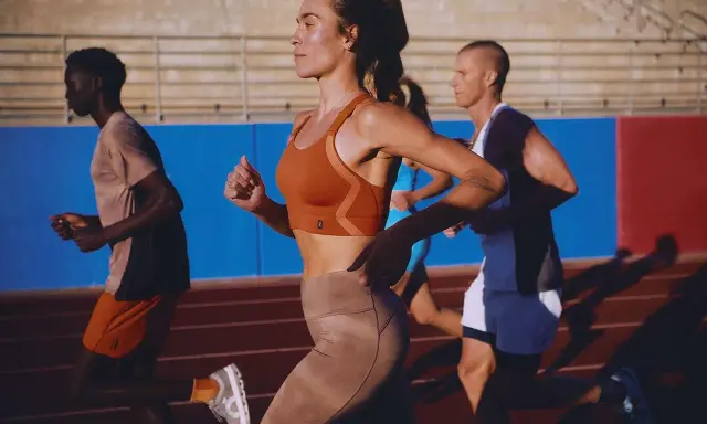 Biegacze na bieżni. Kobieta w pomarańczowym topie i beżowych legginsach, mężczyzna w koszulce i szortach. Buty sportowe z niskim dropem zapewniają komfort.