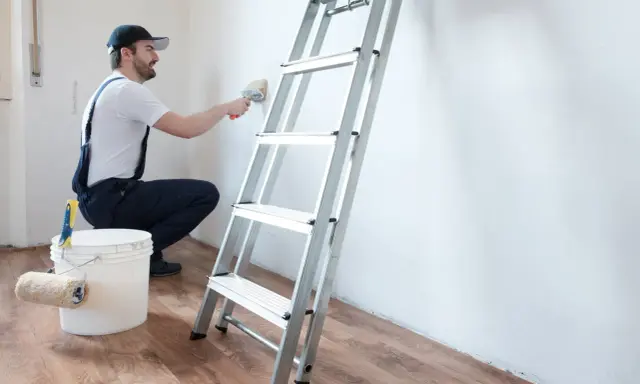 Hombre con mono azul y gorra usa brocha para limpiar paredes, junto a escalera y cubo con rodillo.