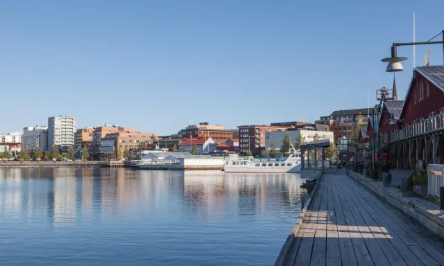 En vacker dag att besöka Luleå. En träspång leder längs vattnet förbi röda byggnader och en vit båt.