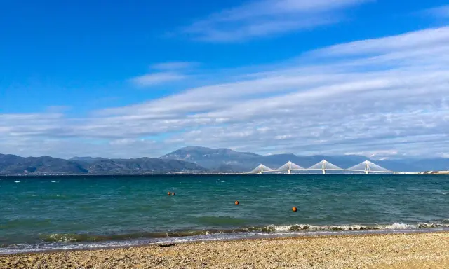 Spokojne morze i plaża w Grecji w styczniu. W oddali widać most Rio-Antirio i góry.