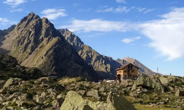Il rifugio Genova, incastonato tra imponenti montagne rocciose e un cielo azzurro punteggiato di nuvole, offre un riparo solitario e suggestivo.