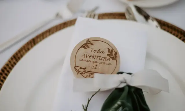 Detalle de mesa de boda con un distintivo de madera que dice "Toda aventura comienza con un SÍ". Ideas para nombres de mesas de boda.