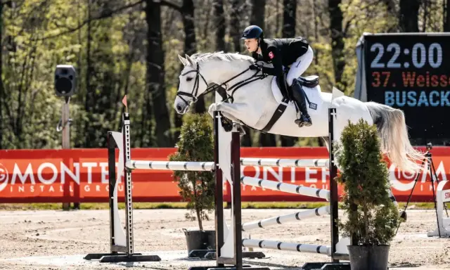 Greta Busacker und ihr weißes Pferd überwinden eine Hürde. Im Hintergrund eine digitale Anzeigetafel.